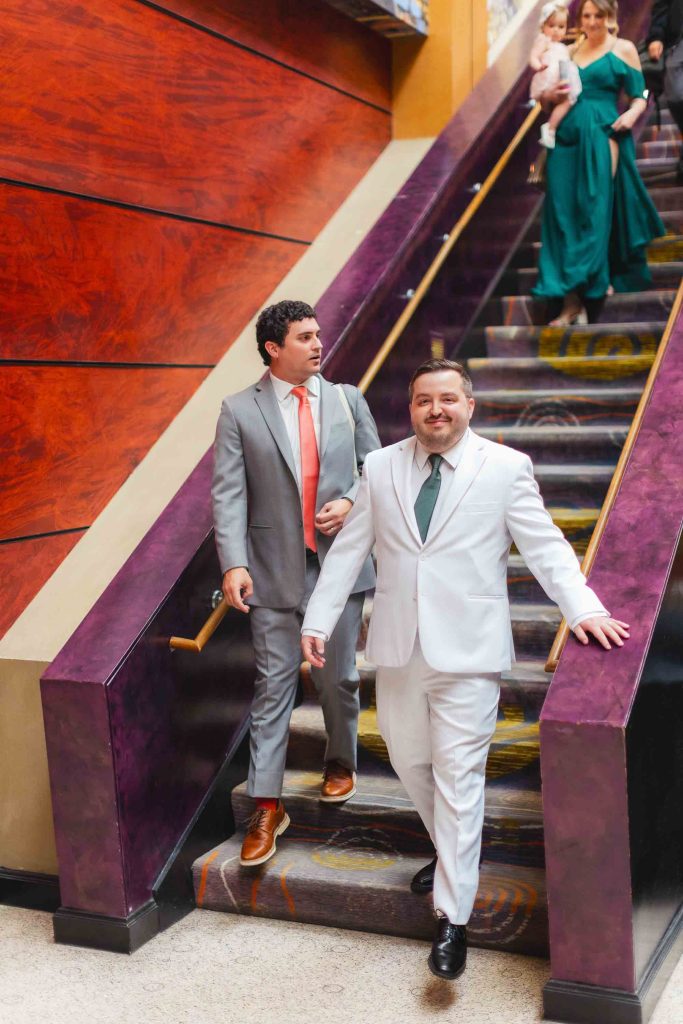 Two men in suits descend indoor stairs, heading to a Pier 5 Hotel wedding first look—one in a light gray suit with an orange tie, the other in white with a green tie. Two women and a child are visible in the background at the top of the stairs.