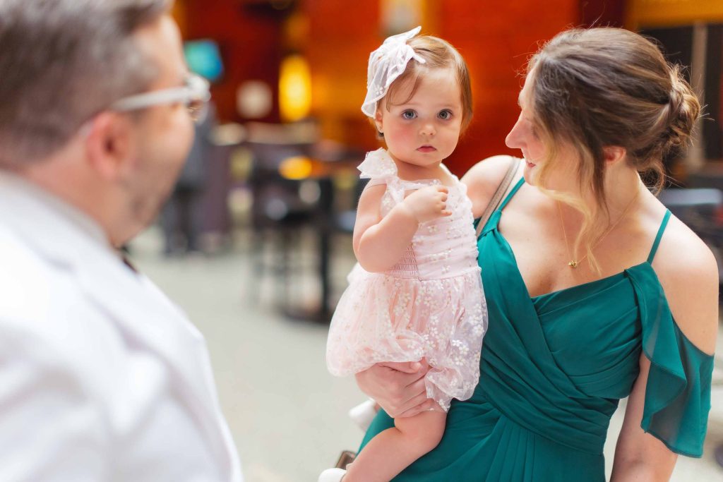 A woman in a green dress holds a young girl in a pink dress and bow while sharing a heartfelt moment with a man in a white jacket during a wedding First Look at Pier 5 Hotel.