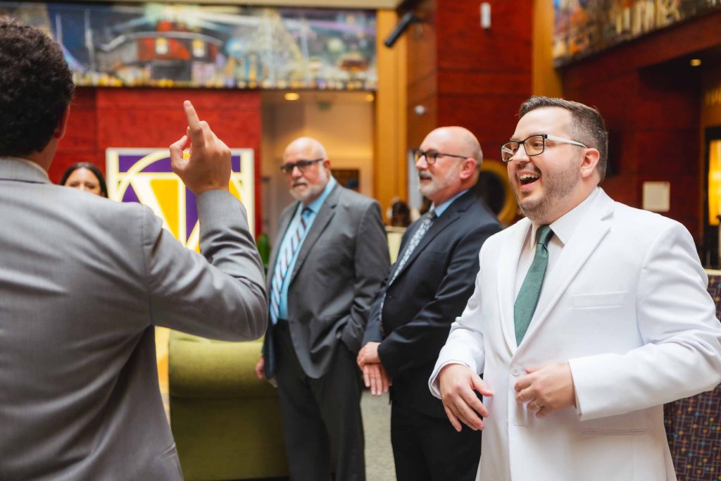 A group of men in suits, one in a white suit, stand indoors at the Pier 5 Hotel having a lively conversation; one person is gesturing with his hand during the wedding's First Look.
