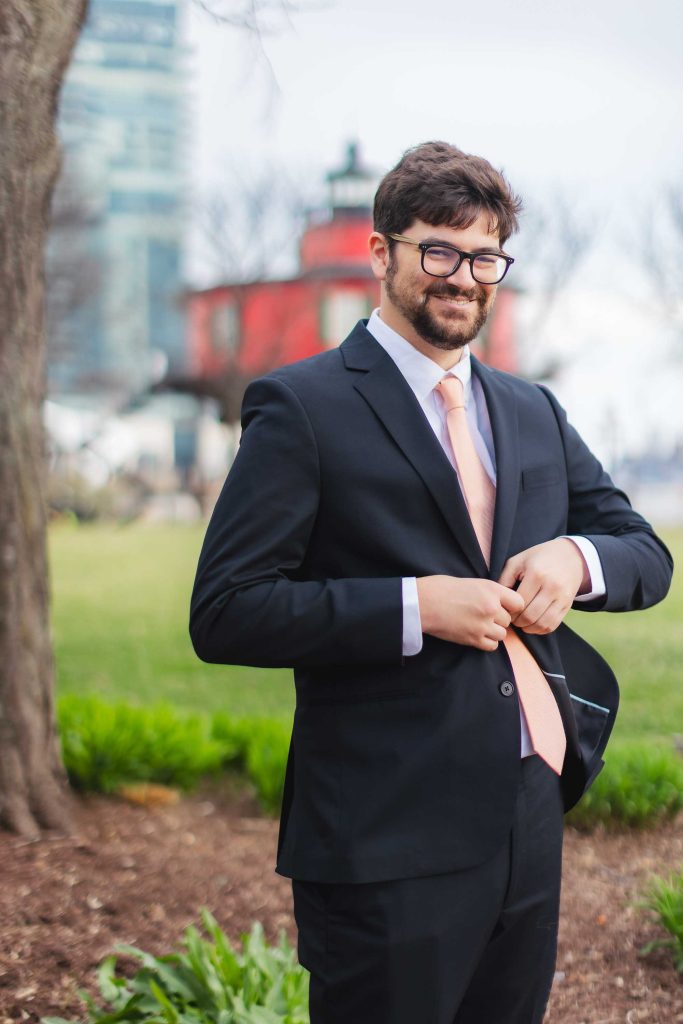 A man in a black suit and peach tie stands outdoors at Pier 5 Hotel, buttoning his jacket during a First Look, with a red building and greenery in the background.