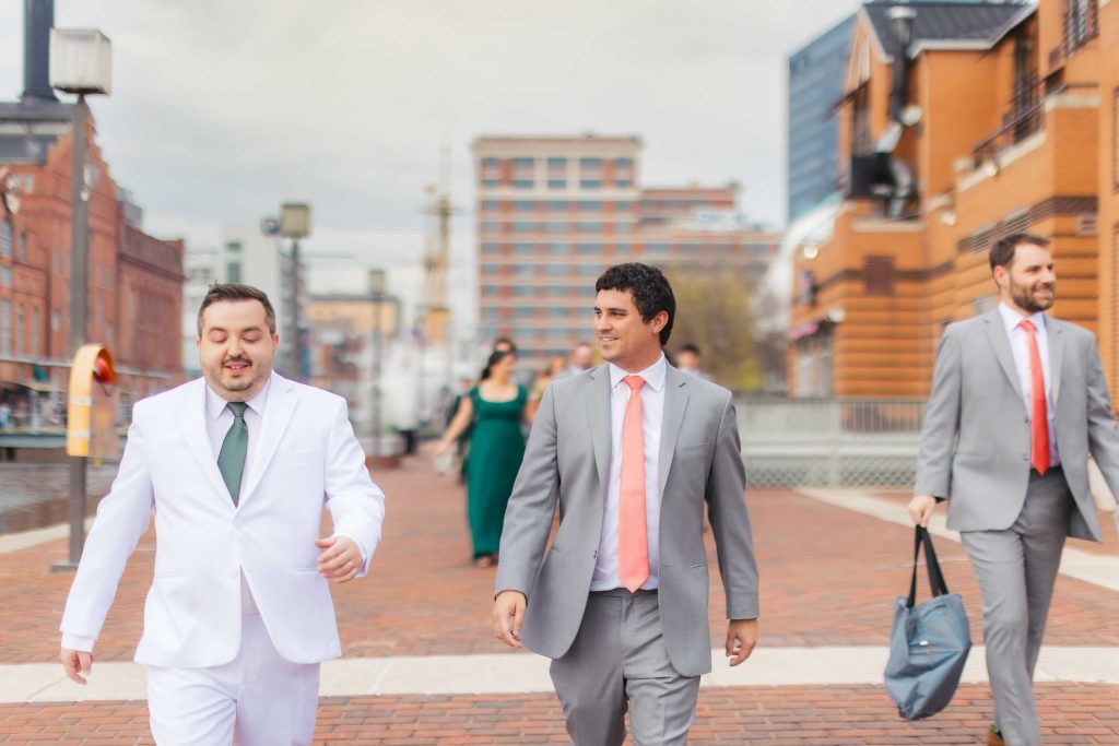 Three men in suits, one in white and two in gray, walk outside on a brick pathway near Pier 5 Hotel for a Wedding First Look, with city buildings and other people in the background.