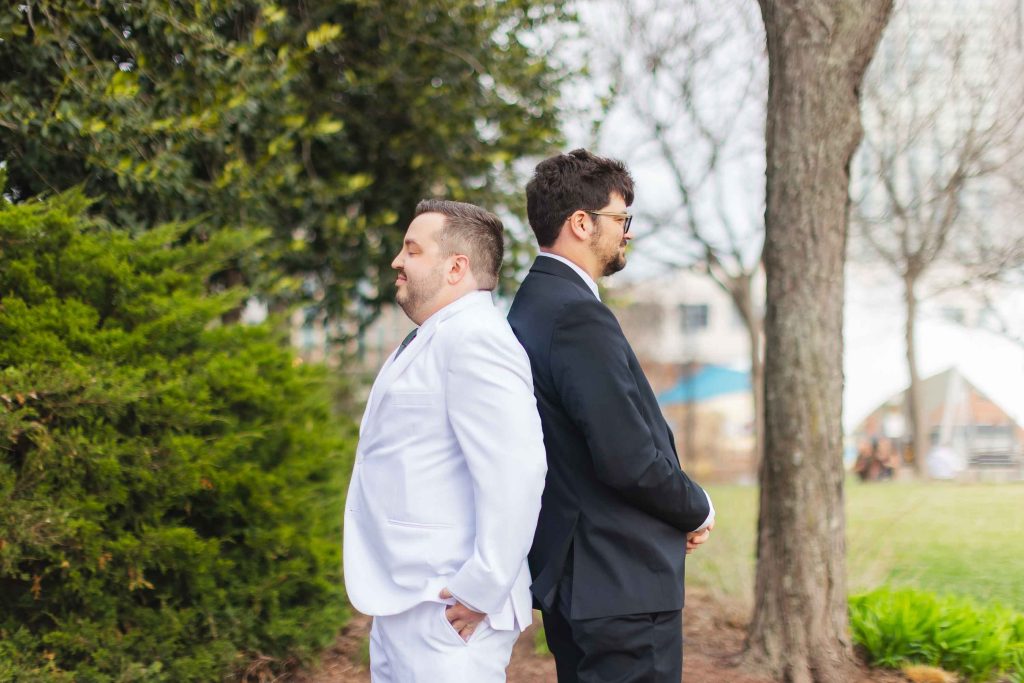 Two men stand back to back outdoors during a wedding first look; one wears a white suit, the other a black suit. Trees and greenery surround them, with the Pier 5 Hotel in the background.