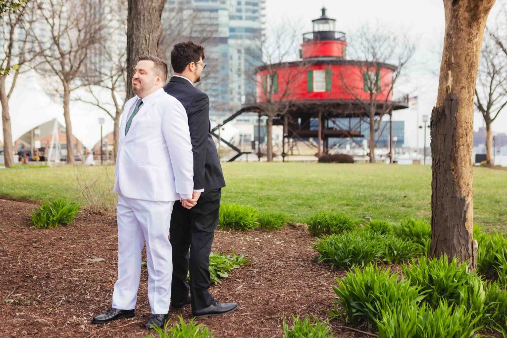 Two men in suits, one in white and one in black, share a First Look back-to-back, holding hands in a park near Baltimore’s Pier 5 Hotel, with a red lighthouse and city buildings as their backdrop.
