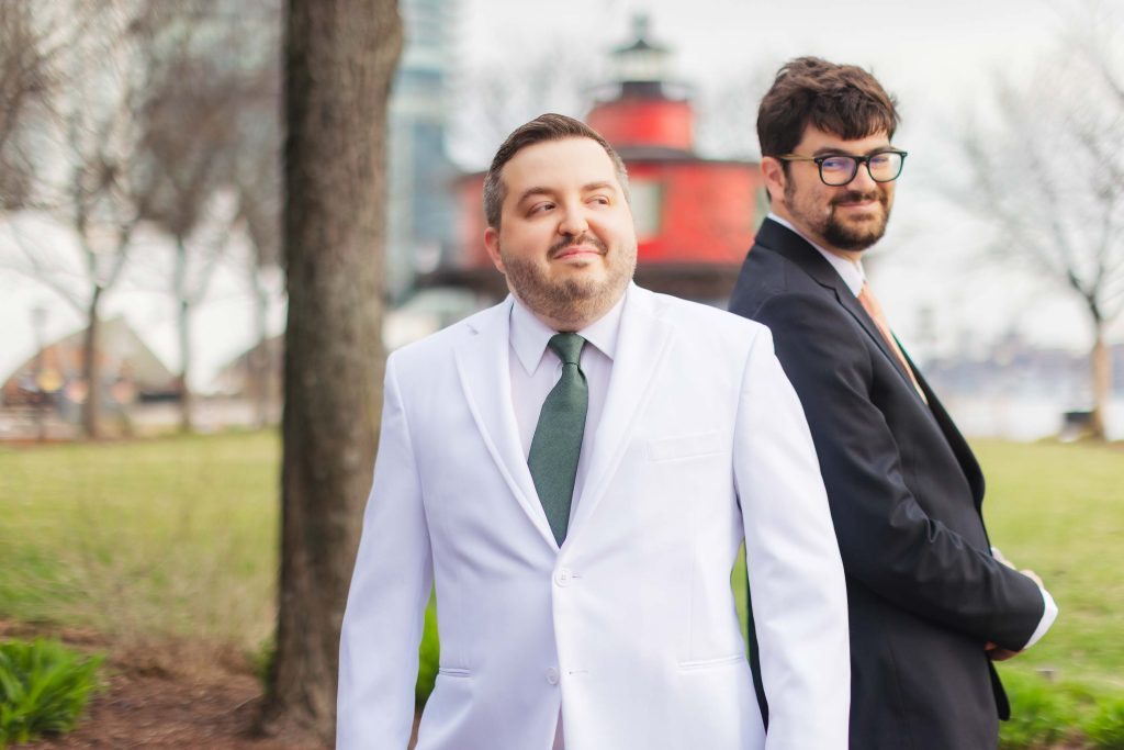 Two men in suits, one in white and one in black, stand back to back outdoors near a red lighthouse and trees at Pier 5 Hotel, sharing a special First Look on their wedding day under cloudy skies.