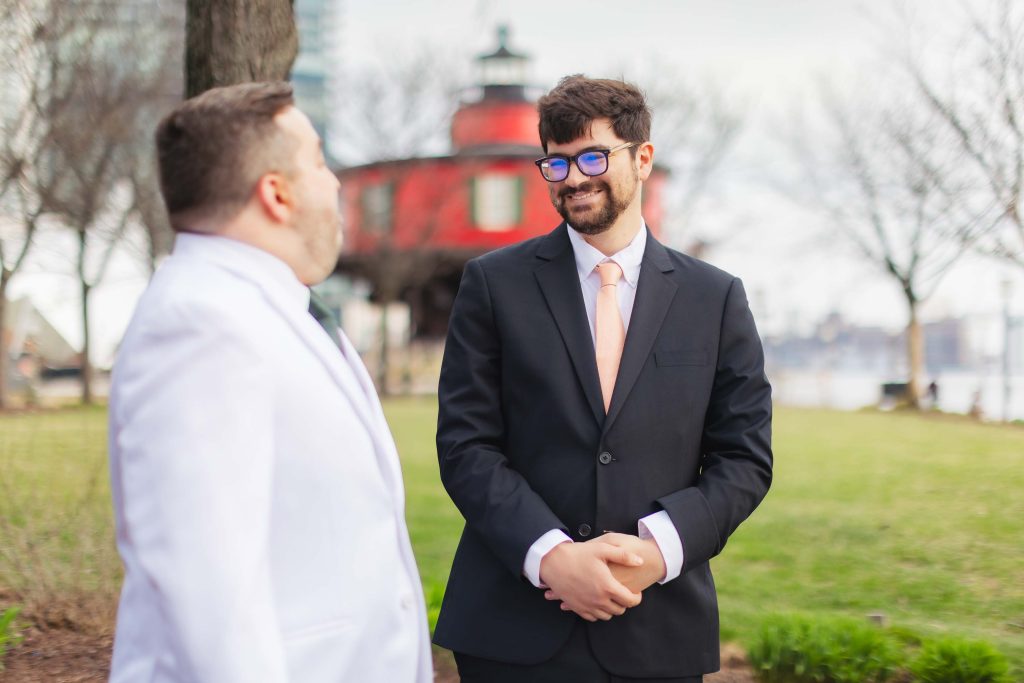 Two men in suits stand outdoors near a red lighthouse at Pier 5 Hotel; one wears a white suit, the other a black suit with a peach tie, as they share a conversation during a wedding First Look moment.
