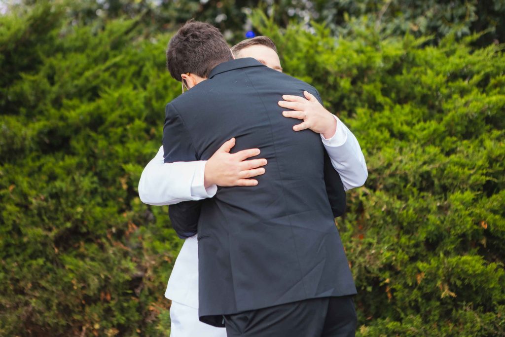 Two people wearing suits, one in black and one in white, are hugging outdoors in front of green bushes—capturing a heartfelt moment at a wedding near Baltimore’s Pier 5 Hotel.