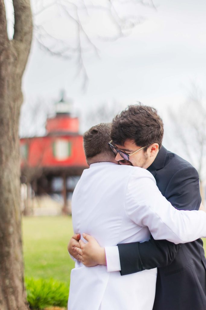 Two men, one in a white suit and one in a black suit, share a wedding embrace outdoors near a tree, with the Pier 5 Hotel's red building visible in the background.