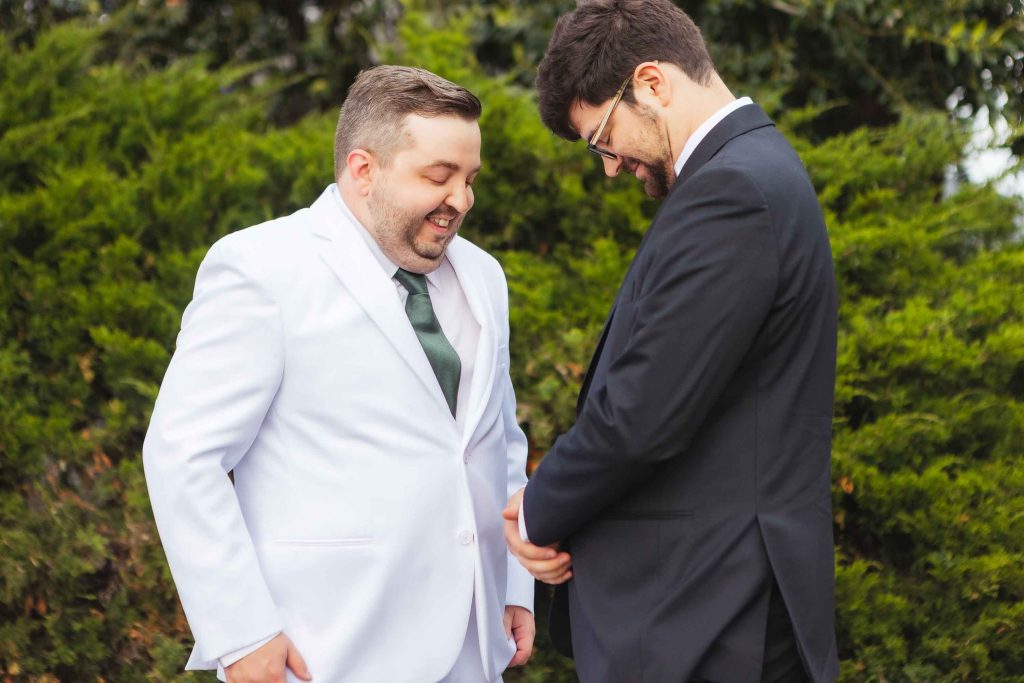 Two men in suits, one in white and one in black, share a joyful First Look outdoors at Pier 5 Hotel in Baltimore, smiling and gazing down with green bushes behind them.