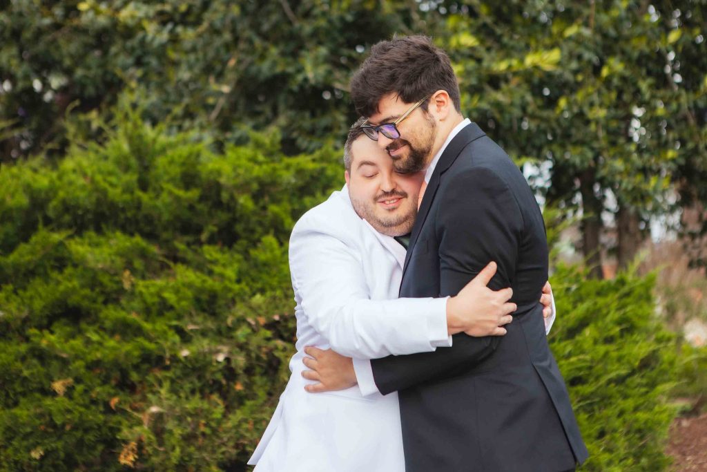 Two men in suits—one in white, one in black—share a heartfelt hug outdoors in front of green bushes and trees, capturing a touching First Look moment on their wedding day in Baltimore.