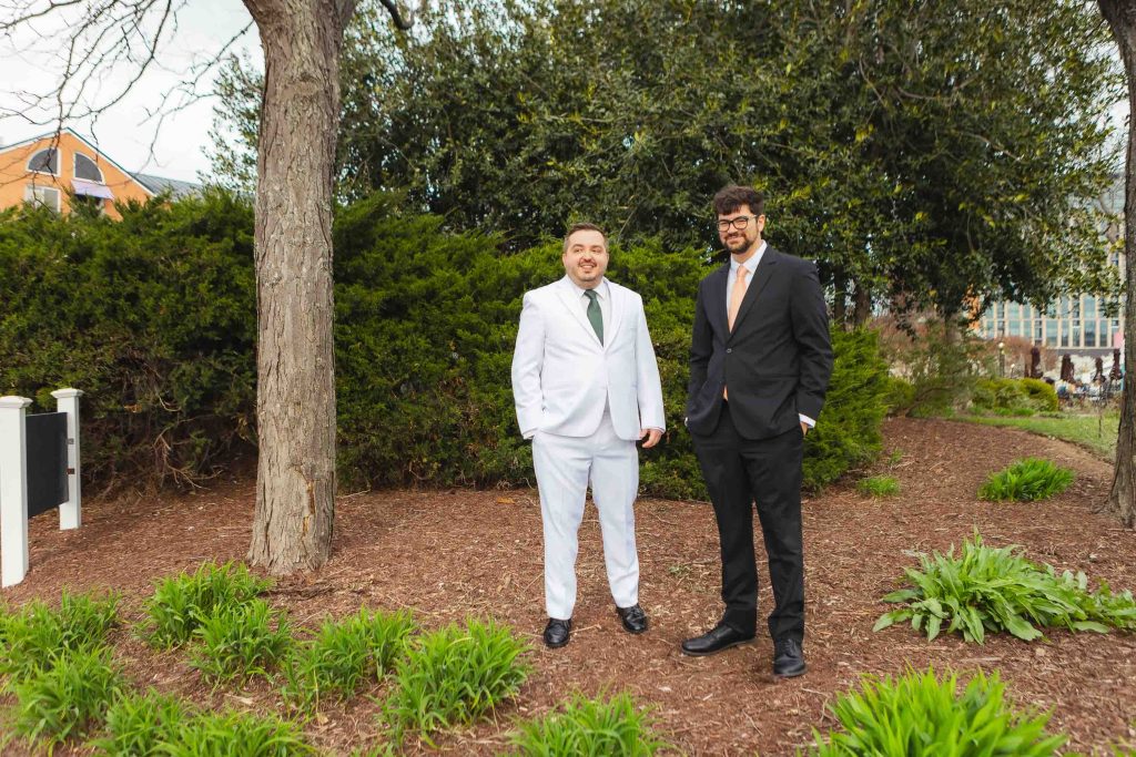 Two men share a First Look outdoors on mulch near green plants and trees at Pier 5 Hotel in Baltimore—one in a white suit with a green tie, the other in a black suit with a peach tie.