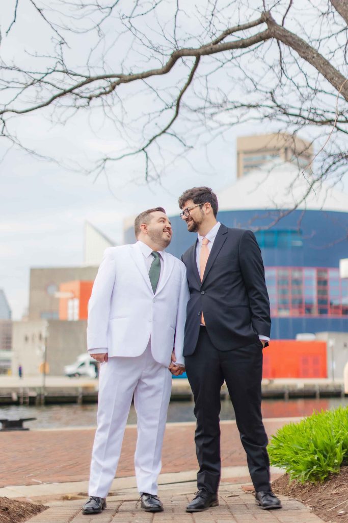 Two men in suits, one in white and one in black, stand close together outdoors on a brick path at Pier 5 Hotel, holding hands and smiling at each other during their wedding first look.