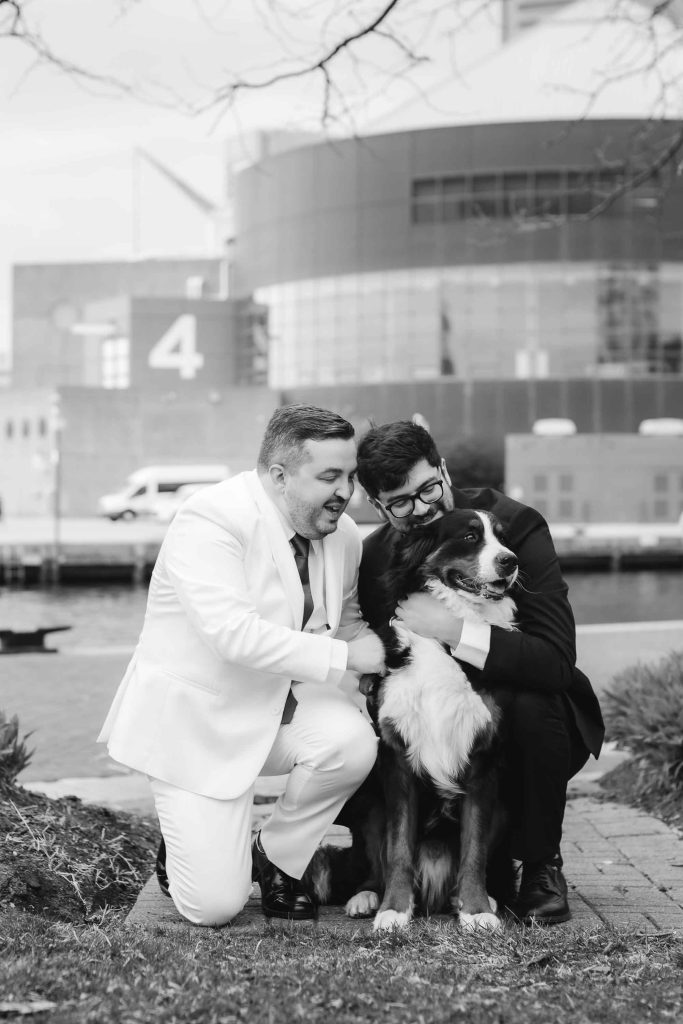 Two men in suits, one white and one black, kneel outdoors next to a large dog near the National Aquarium in Baltimore, with a modern building and water in the background.