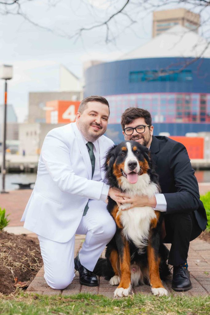 Two men, one in a white suit and one in black, kneel outdoors beside a large Bernese Mountain Dog. The National Aquarium and water set the scene in Baltimore, creating striking portraits with modern architecture as the backdrop.