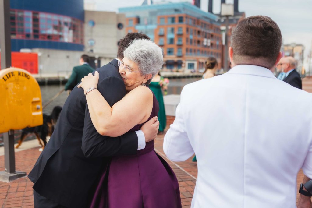 An older woman in a purple dress hugs a man in a dark suit outdoors during a First Look at Baltimore’s Pier 5 Hotel, with other people and city buildings visible in the background.