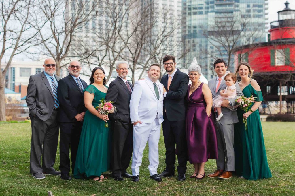 A group of eleven people dressed formally, including a bride holding a child, pose outdoors on grass in Baltimore with city buildings and the National Aquarium’s red structure in the background.