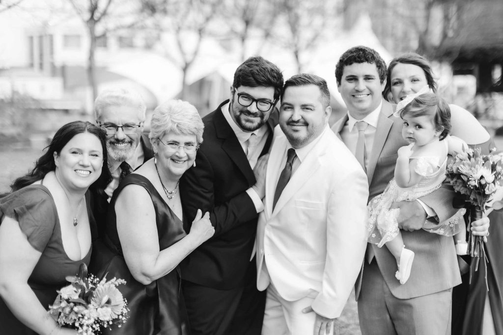 A group of eight adults and one child, dressed formally, pose closely together and smile for a photo outdoors at a wedding in Baltimore.
