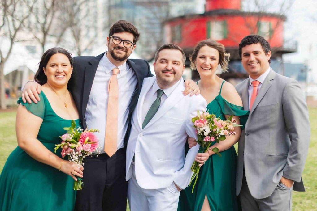 Five people dressed in formal attire pose outside the National Aquarium in Baltimore. The group, attending a wedding, includes three men in suits and two women in green dresses, smiling at the camera while two hold bouquets.