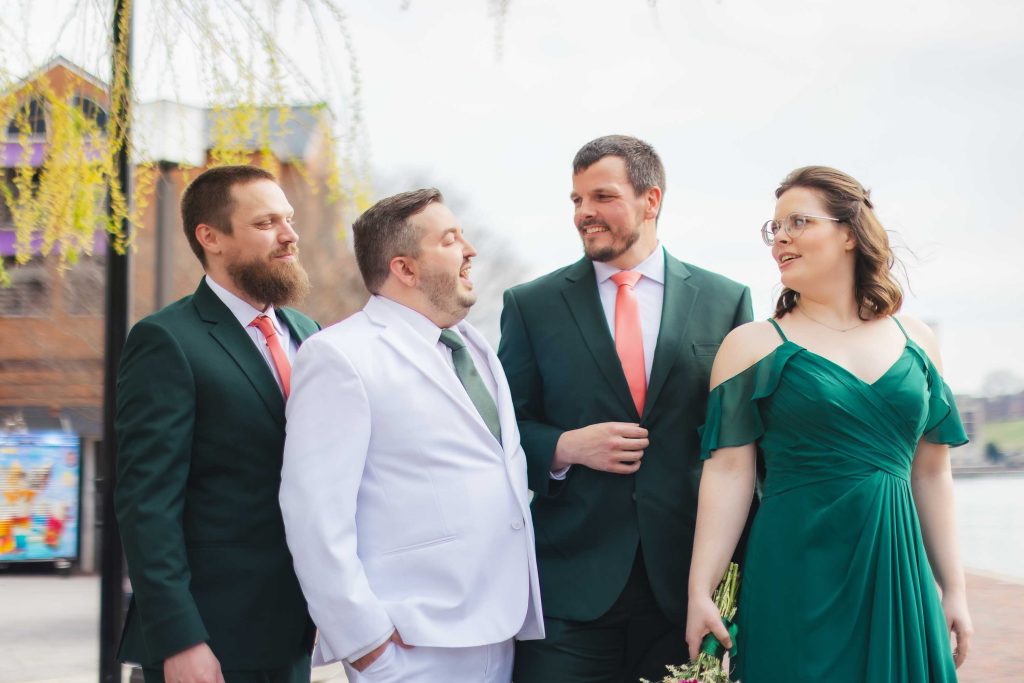 Four people in formal attire stand outdoors, smiling and talking as if posing for wedding portraits. Three wear dark green, one a white suit, with a bouquet in hand.