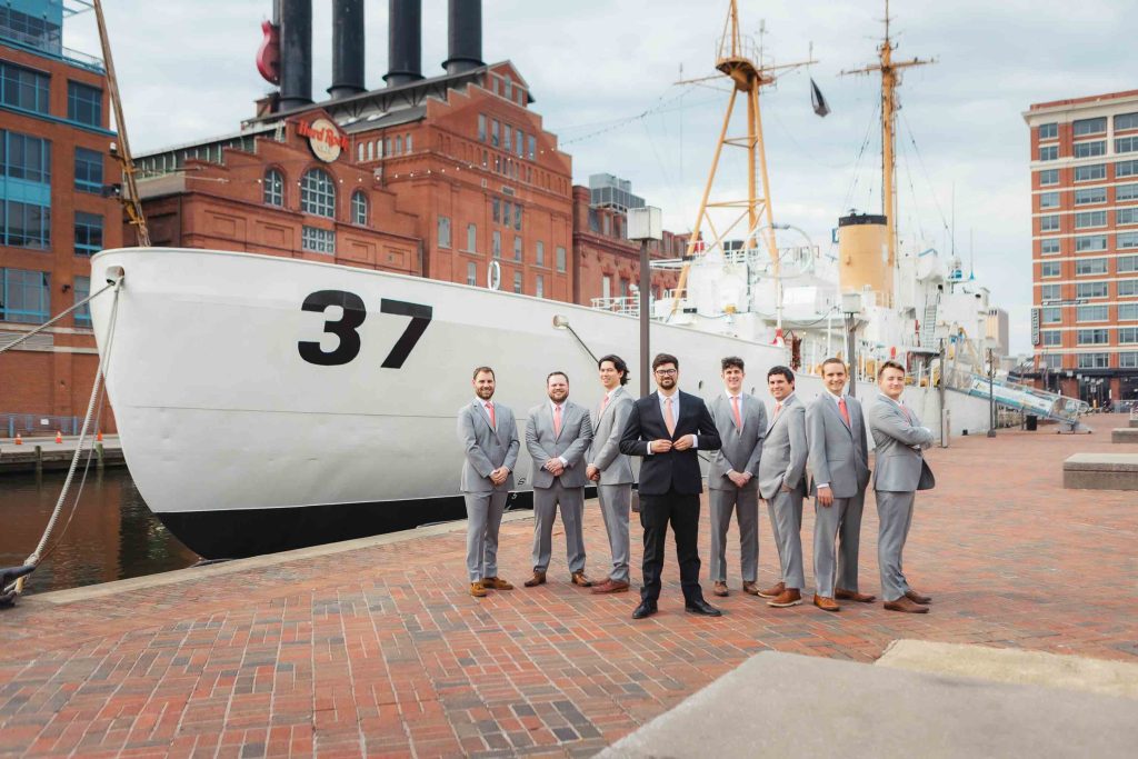 Eight men in suits stand on a brick walkway in front of a white ship with the number 37, surrounded by Baltimore city buildings, near the National Aquarium—perfect for a wedding photo op.
