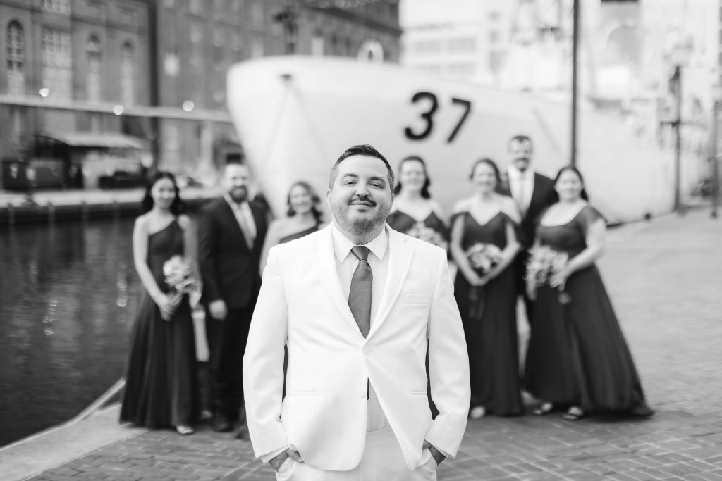 A man in a white suit stands in front of a group of people in formal attire by a docked ship marked with the number 37 in Baltimore. The black and white image resembles vintage portraits by the harbor.