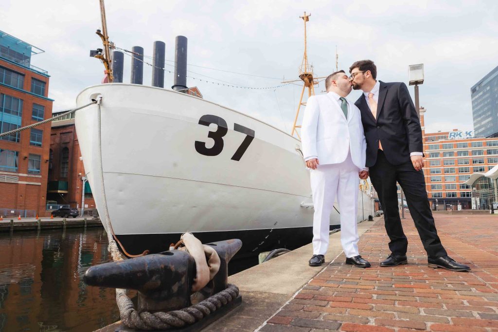 Two men in suits hold hands and kiss on a dock beside a large white ship marked 37, with the National Aquarium and city skyline in the background—capturing heartfelt wedding portraits in an urban harbor setting.