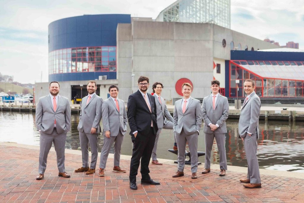 Eight people pose in suits by a waterfront in Baltimore, with modern buildings and the National Aquarium in the background. One wears a black suit while the others wear gray suits with coral ties.