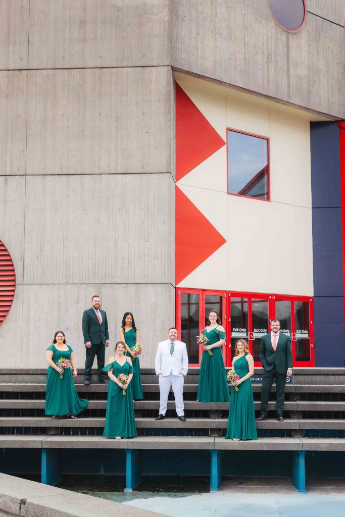 A wedding party poses on concrete steps in front of a modern building with geometric designs in Baltimore. Four women in green dresses, three men in suits, and one man in a white suit hold bouquets for group portraits.