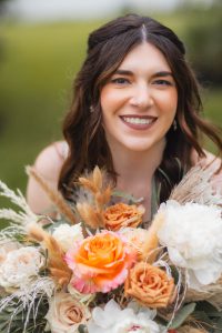 A smiling woman holds a floral bouquet containing orange, pink, and white flowers. She is outdoors at a picturesque vineyard, with lush greenery in the background.