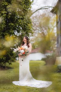 A bride in a white wedding dress stands outside, holding a large bouquet of flowers. The background features the lush greenery of The Barns at Hamilton Station vineyard and part of a charming building.
