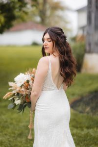 A bride with long, wavy hair stands outdoors in a vineyard, facing slightly to the side, holding a bouquet with white and beige flowers. She is wearing a white, sleeveless lace wedding dress with an open back.