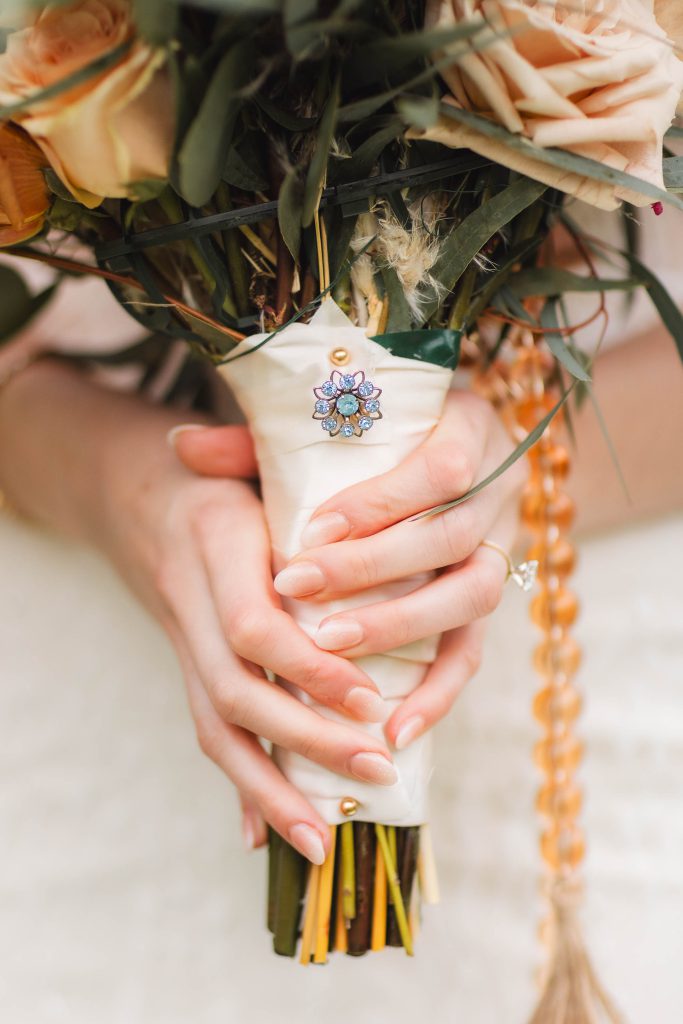 Close-up of hands holding a bouquet of flowers wrapped in white fabric, adorned with a jeweled brooch. The hands have neatly manicured nails, and one wears a diamond ring. The intricate details make it perfect for a wedding at The Barns at Hamilton Station.