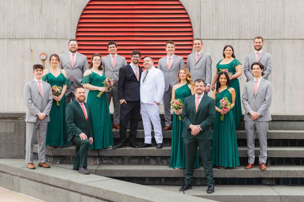 A wedding party poses on concrete steps outside the National Aquarium in Baltimore; the group includes people in green dresses, grey suits with pink ties, and one person in a white suit with a red tie, against a modern building background.