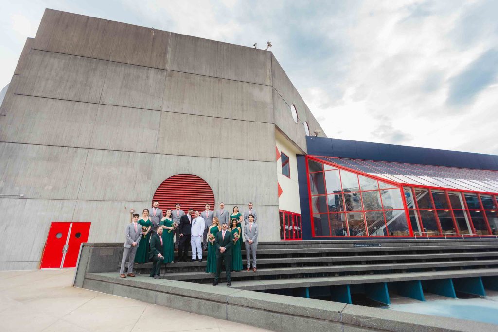 A group of people, some in graduation gowns, stand on steps outside a modern concrete building with red and glass architectural features in Baltimore.