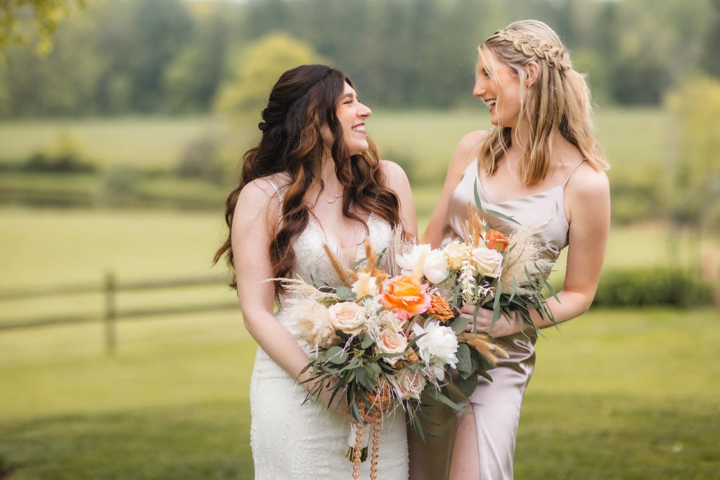 Two women smiling at each other outdoors, each holding a bouquet of flowers. One is wearing a white dress, and the other is in a light-colored dress. A blurred green field in the background hints at a picturesque vineyard, creating an enchanting wedding portrait.