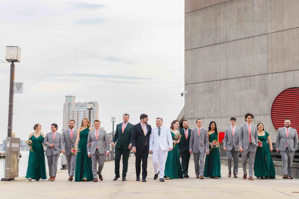 A wedding party poses outdoors in Baltimore; the grooms stand in the center, surrounded by attendants in gray suits with pink ties and green dresses, with cityscape portraits and the National Aquarium in the background.