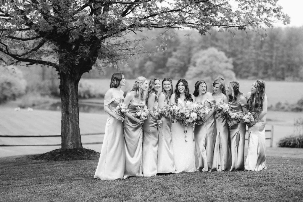 A group of bridesmaids in matching light-colored dresses hold bouquets and smile together under a tree in an outdoor setting, creating the perfect wedding portrait at The Barns at Hamilton Station.