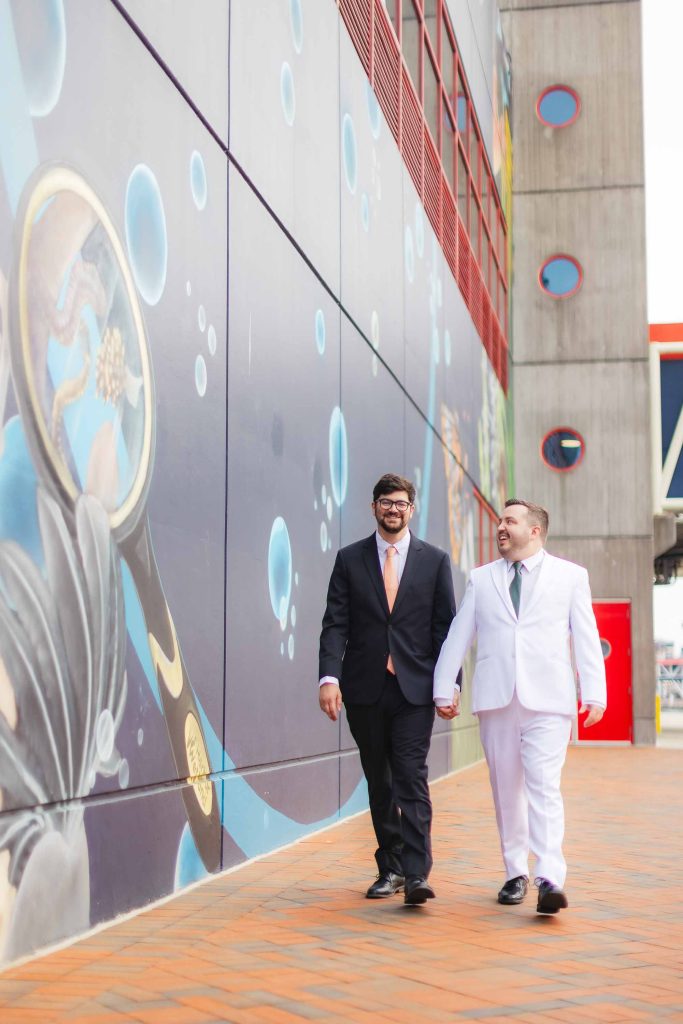 Two men in suits, one black and one white, hold hands and walk beside a colorful mural on an urban sidewalk—capturing stylish portraits reminiscent of a wedding day near the vibrant scenes by the National Aquarium.