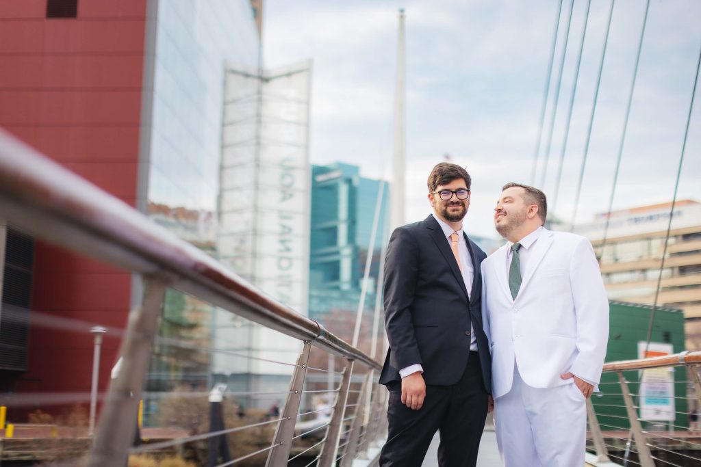 Two men in suits, one in black and one in white, stand together on a modern pedestrian bridge in Baltimore, with glass buildings and the National Aquarium visible in the urban background.