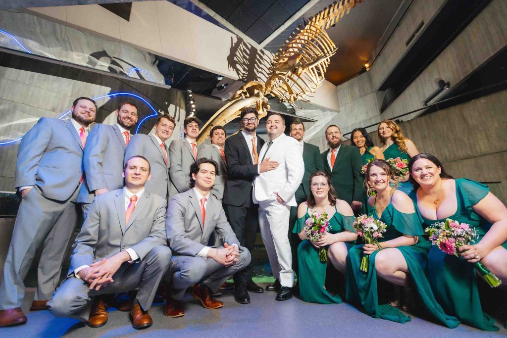 A wedding party poses indoors at the National Aquarium in front of a dinosaur skeleton exhibit; the group includes people in gray suits with orange ties and green dresses, centered around two grooms. Perfect for memorable wedding portraits.