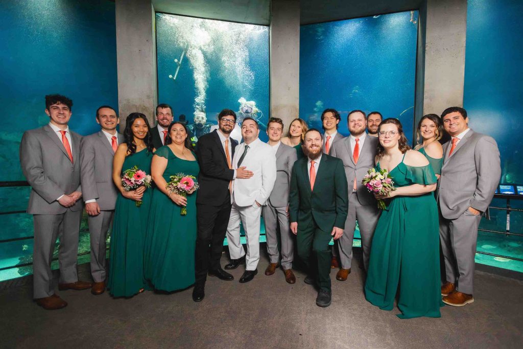 A wedding party poses for portraits indoors at the National Aquarium in Baltimore, with two grooms in white and black suits and attendants in grey suits and green dresses holding bouquets before massive aquarium tanks.