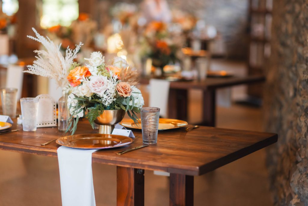 A wooden table is set with gold-rimmed plates, clear glasses, and a flower arrangement featuring white, peach, and orange flowers. The wedding at The Barns at Hamilton Station showcases such elegant details, with the background revealing more similarly decorated tables.