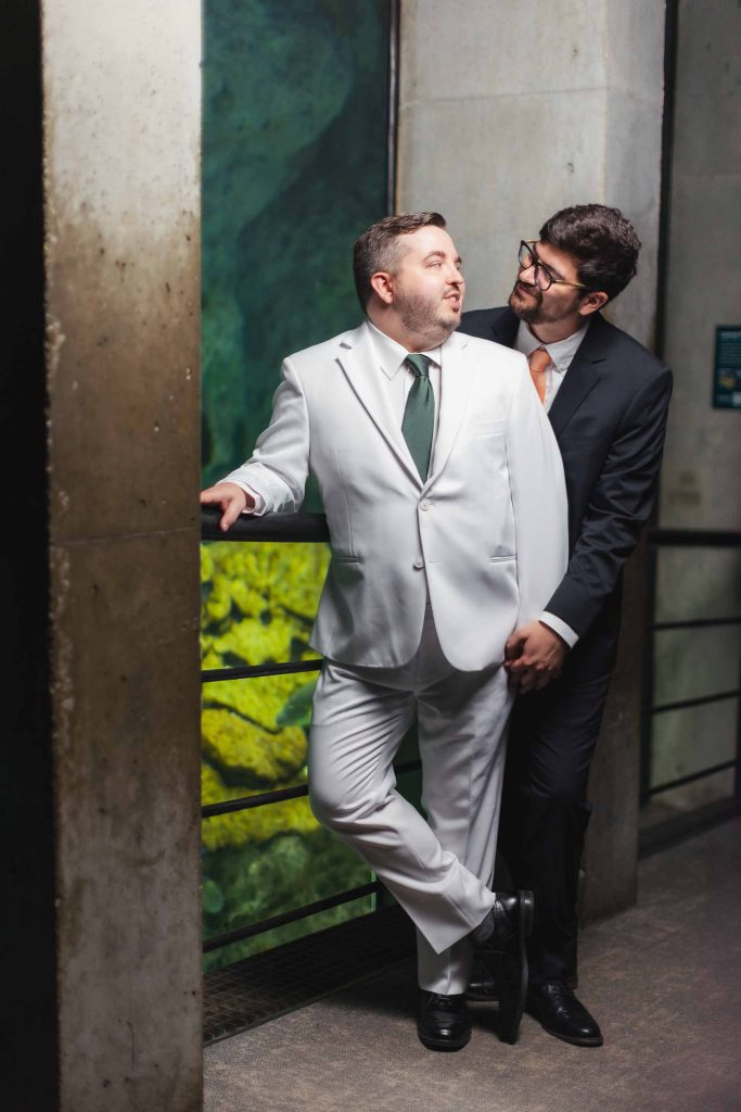 Two men in suits, one in white and one in black, stand closely together near a railing at the National Aquarium in Baltimore, with a large aquarium display behind them—an elegant setting for portraits.