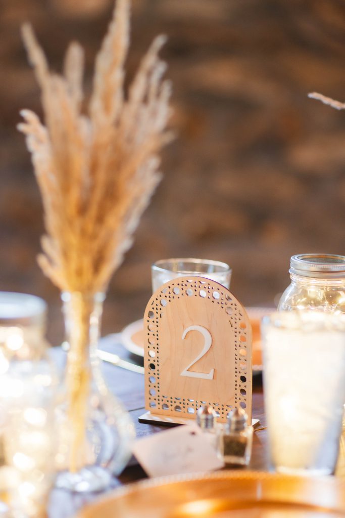 Close-up of a table setting at The Barns at Hamilton Station featuring a decorative table number 2, dried wheat stalks in a vase, and glass jars with string lights, creating a warm and rustic wedding ambiance with charming details.
