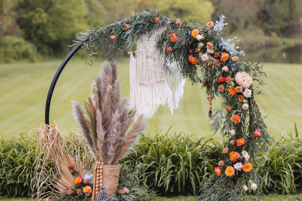 A semi-circular floral arch with greenery, orange and white flowers, and a hanging macrame decor graces the backdrop at The Barns at Hamilton Station. Detailed with a basket of pampas grass and decorated leaves beside it, every element captures the essence of a perfect wedding setting outdoors.
