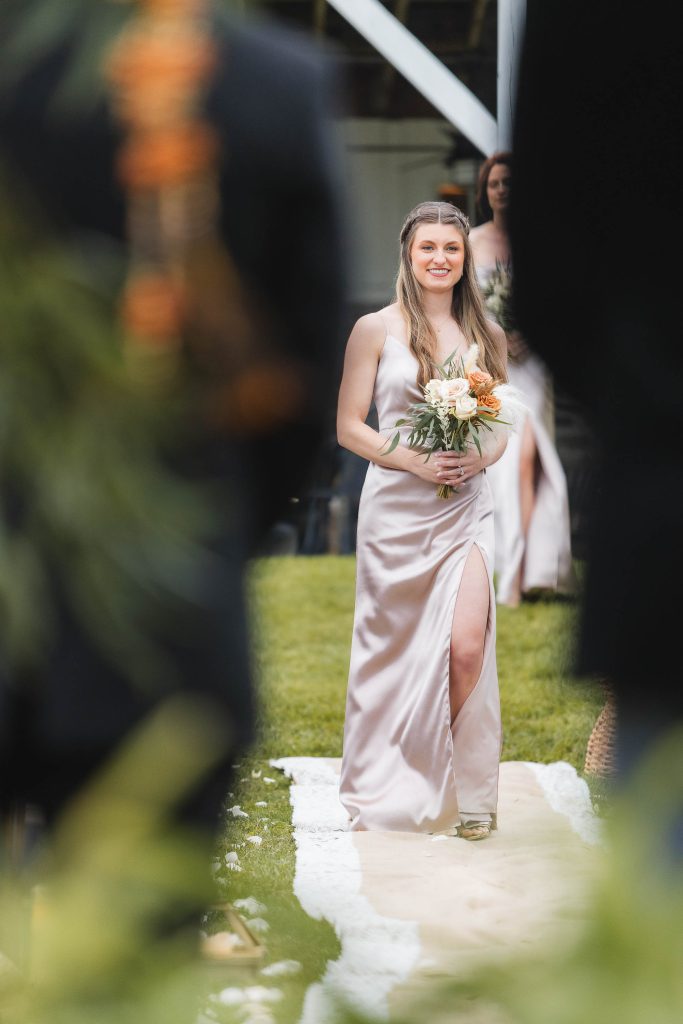 A woman wearing a light-colored dress walks down an outdoor aisle at The Barns at Hamilton Station, holding a bouquet of flowers, during the wedding ceremony.