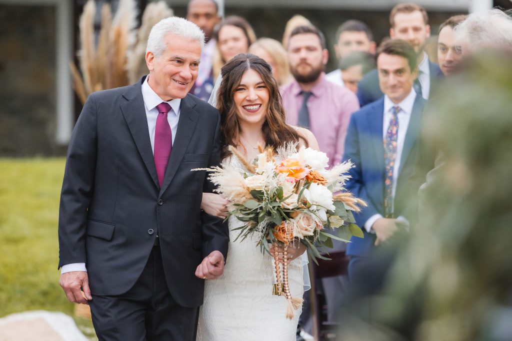 A bride holding a bouquet walks arm-in-arm with a man in a suit down an outdoor aisle at The Barns at Hamilton Station, with guests seated in the background, celebrating this beautiful vineyard wedding.