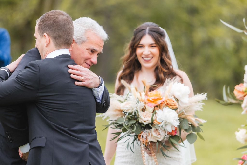 A bride holding a bouquet smiles while a gray-haired man and a younger man in suits hug outdoors at the wedding ceremony, with lush greenery in the background.