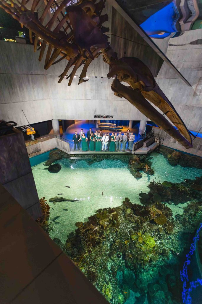 A group of people stands by the edge of a large indoor aquarium at Baltimore’s National Aquarium, with a suspended skeleton model of a marine animal above and various fish and coral visible in the clear water below.