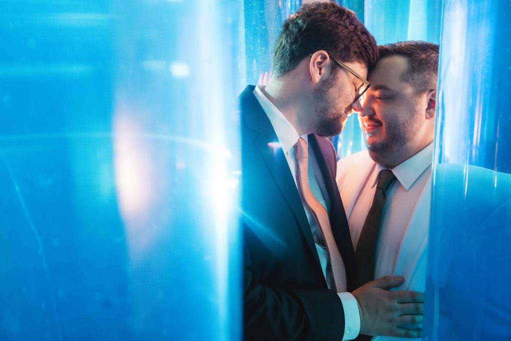 Two men in suits stand close together, smiling and touching foreheads, surrounded by blue light and translucent panels in a scene reminiscent of wedding portraits at the National Aquarium.