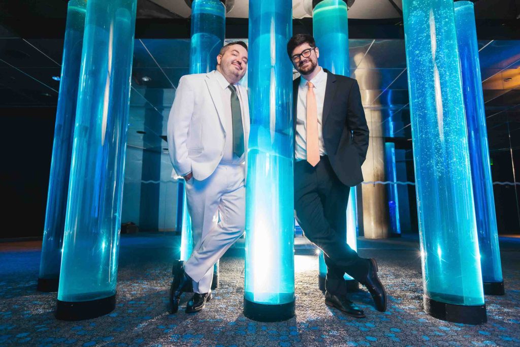 Two men in suits, one in white and one in black, stand and lean against large illuminated blue columns at the National Aquarium in a modern indoor setting.